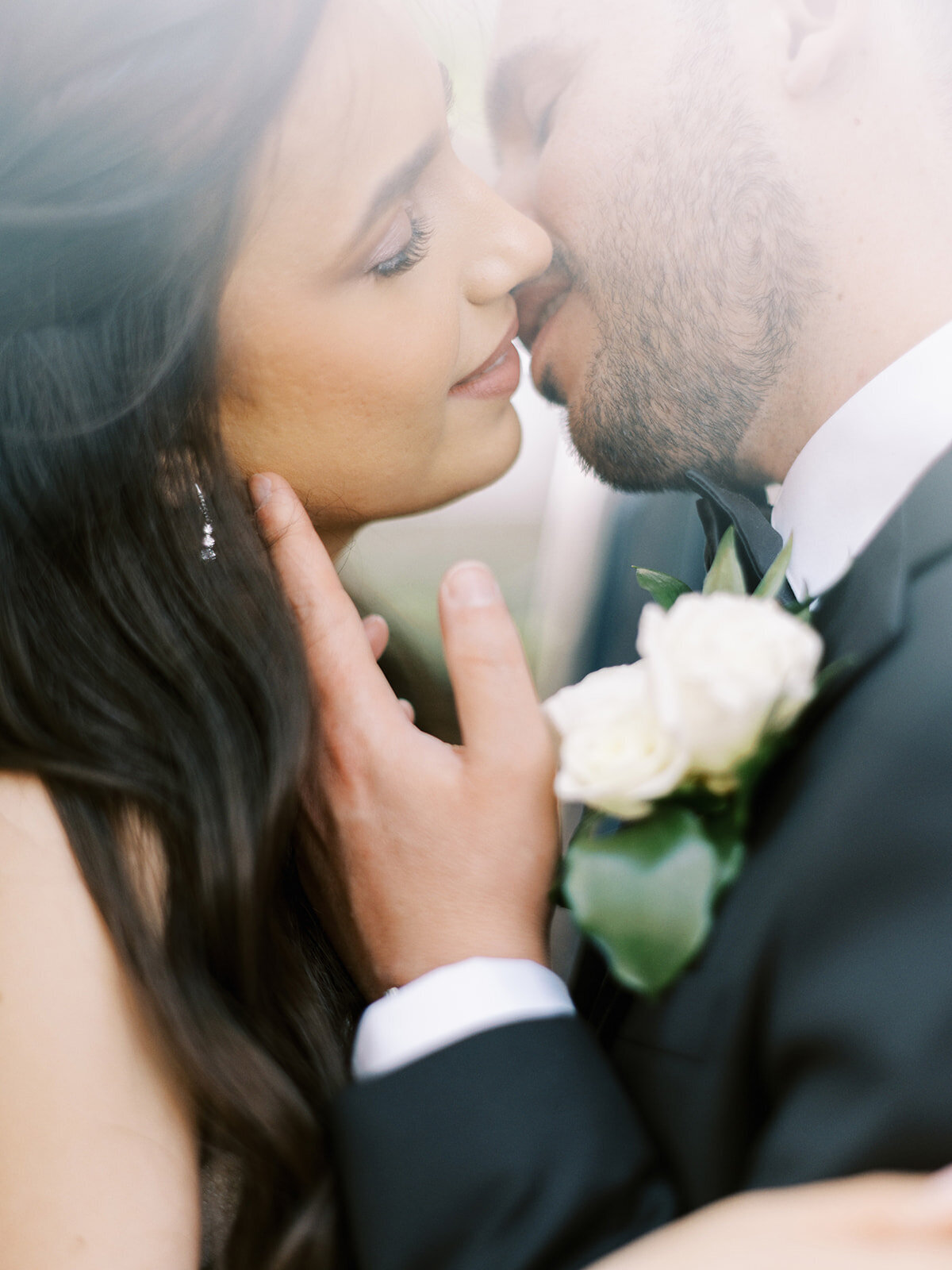 A bride and groom with closed eyes lean in for a kiss, the groom wearing a black suit with a white rose boutonniere, and the bride with long, dark hair and earrings. It’s a classic Calgary wedding set against the elegant backdrop of the Fairmont Palliser.