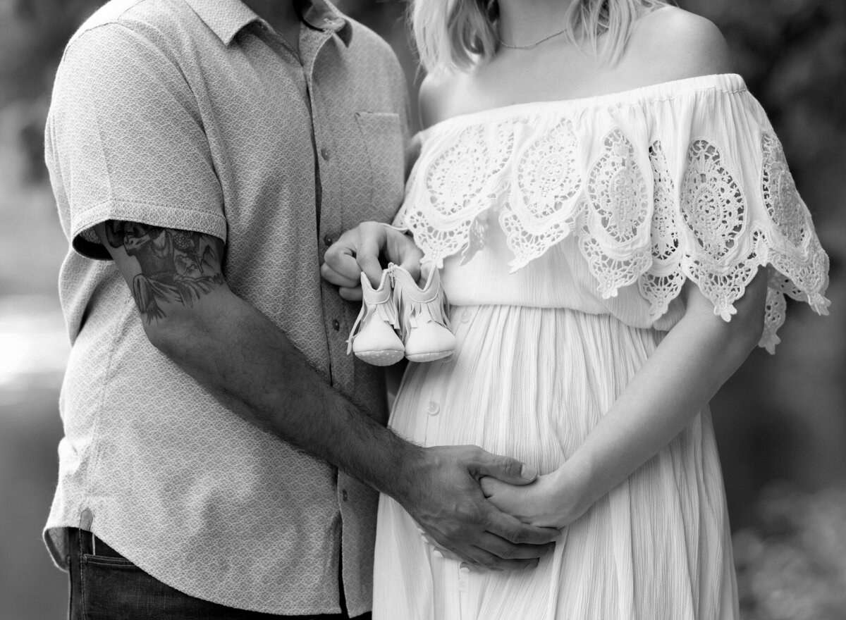 Close-up of hands on baby bump and baby shoes held by couple during Buda, TX maternity shoot