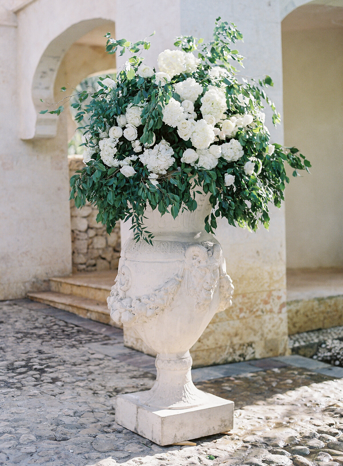 A romantic Caribbean destination wedding scene captured by photographer Laura Gordon. A beautiful bride in a lace wedding dress and her groom in a suit and bow tie, stand closely together in the Dominican Republic. The groom holds the bride's hand as they gaze at each other, bathed in soft sunlight, creating a dreamy tropical ambiance.