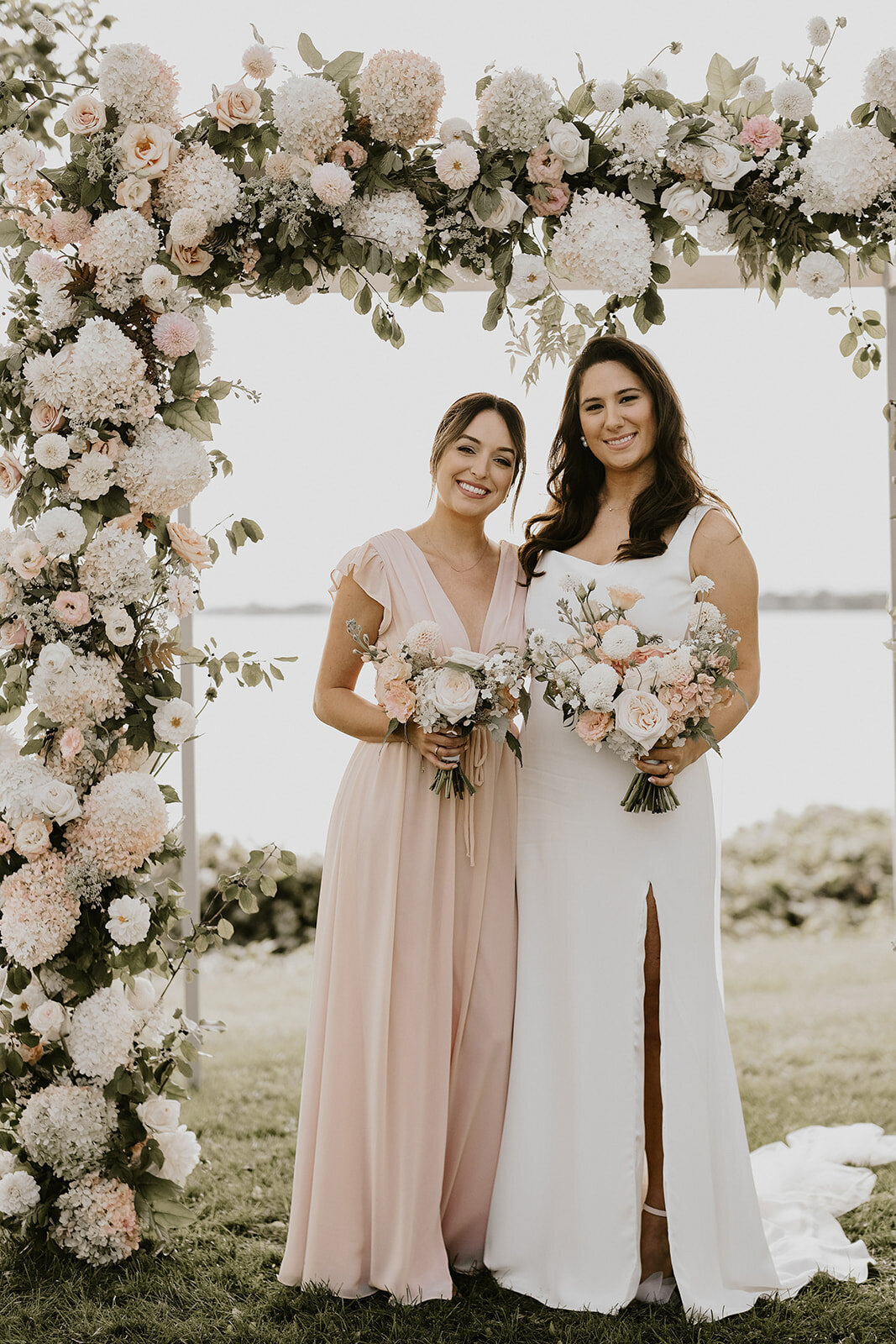 A bride and bridesmaid pose in front of a lush flower ceremony arch at the Willow Inn.