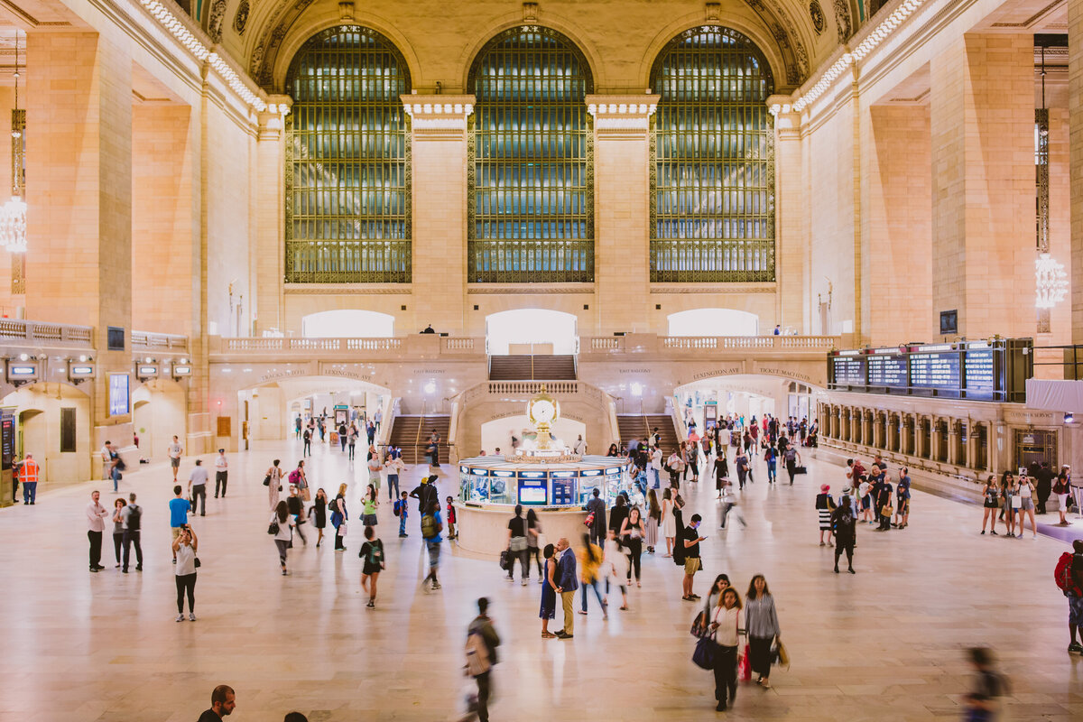 A wide shot of the entire grand central station with an engagement couple standing in the middle sharing a moment with people walking around