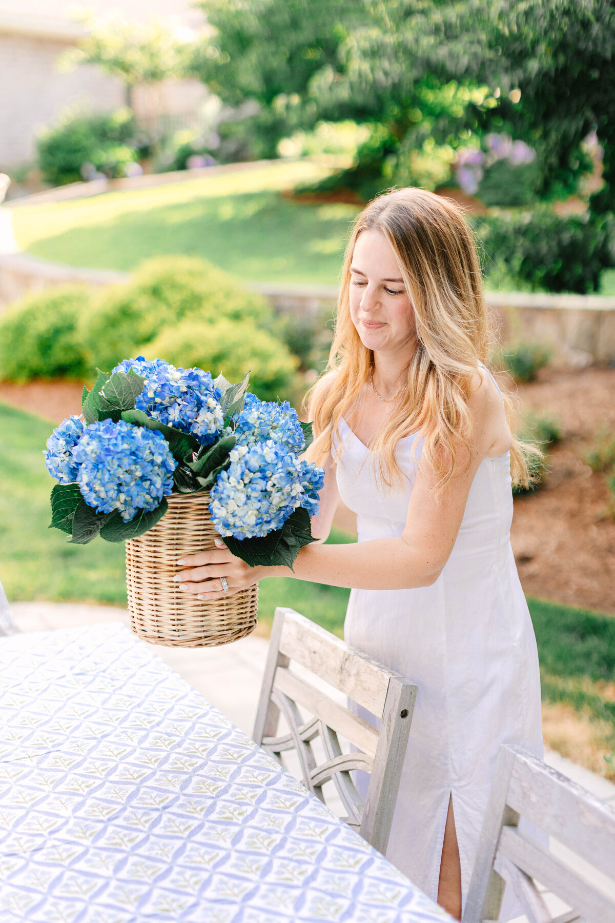 A woman in a white sundress places a wicker basket filled with blue hydrangeas on an outdoor dining table covered with a patterned tablecloth. The lush garden setting in the background adds to the serene atmosphere.