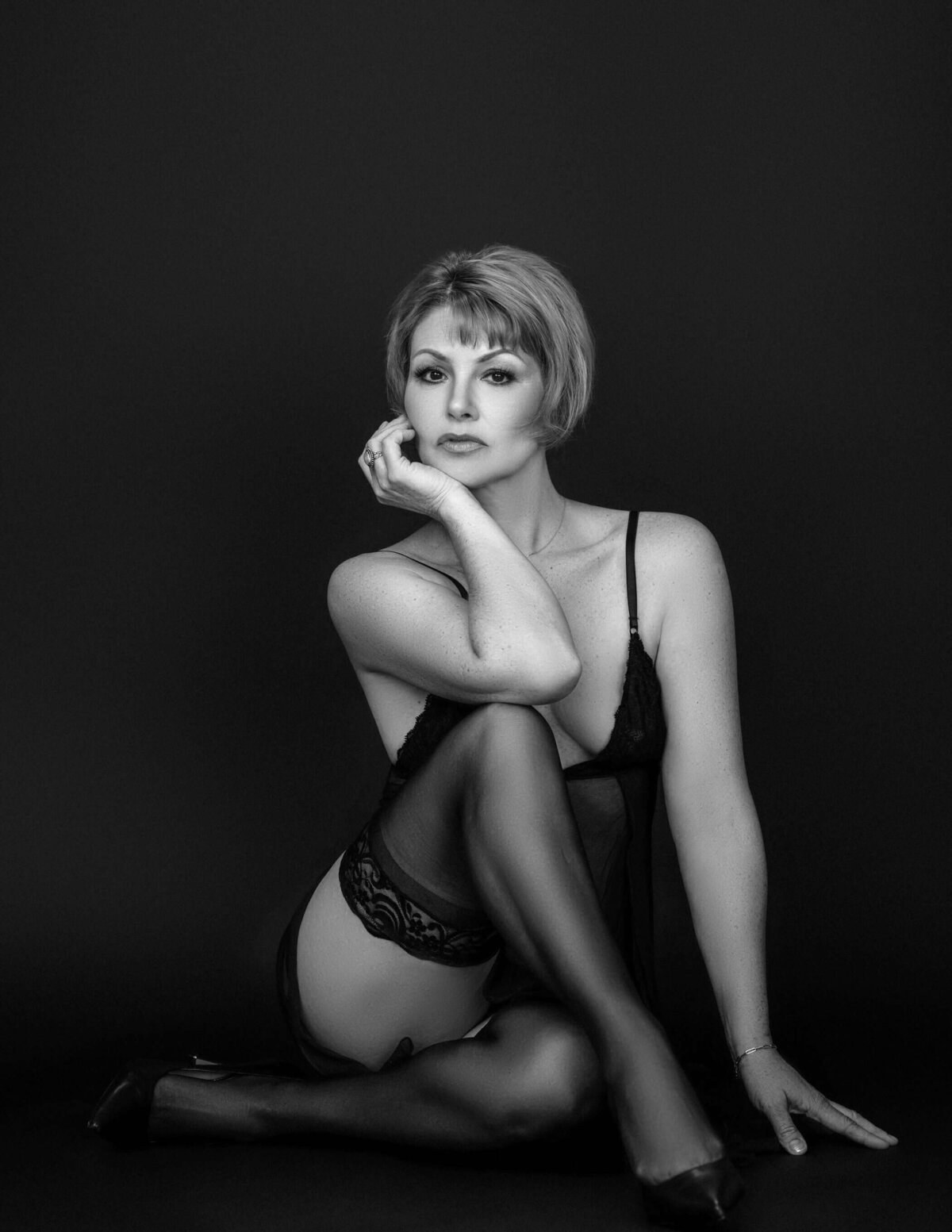 A confident woman in a black sheer lingerie bodysuit and stockings poses on a dark background, exuding elegance and sensuality.