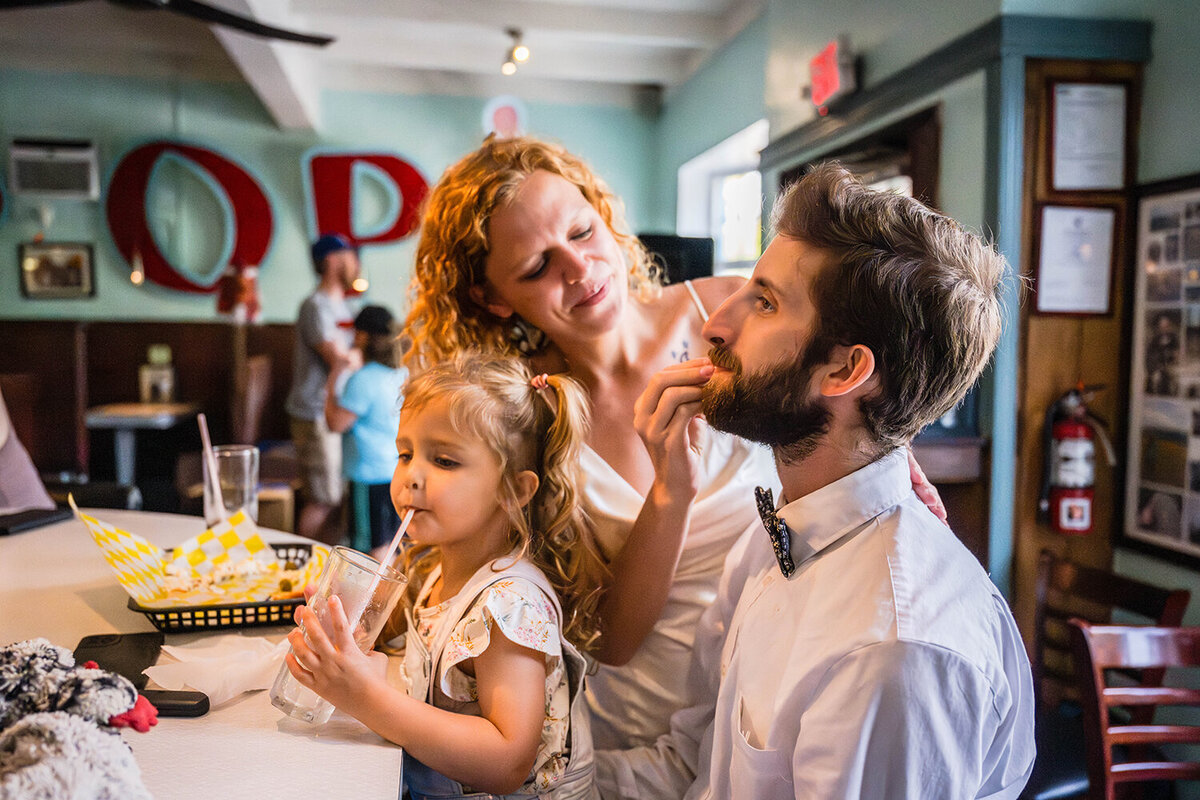 A toddler sips a glass of water through a straw while sitting in her father’s lap. The father is showing his chin to his partner who is wiping off his face and smiling. The family is sitting at the bar of Pop’s Ice Cream and Soda Bar in Grandin Village in Roanoke, Virginia.