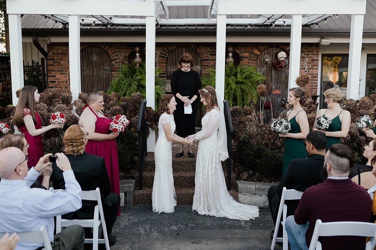 Two brides in lace wedding dresses hold hands in front of their officiant as they exchange wedding vows. The bridesmaids dressed in red and green dresses holding paper flower bouquets stand on either side of them.