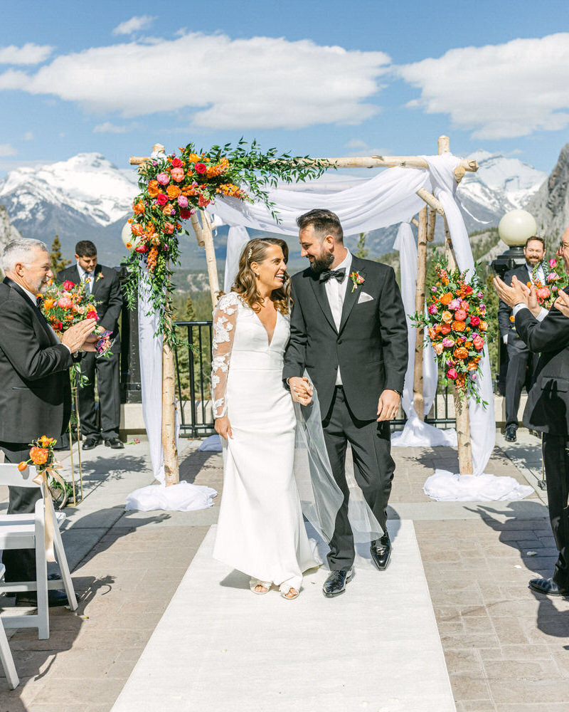 A breathtaking summer wedding at Chateau Lake Louise, where the bride and groom walk down the aisle with the majestic Victoria Glacier and Lake Louise as their backdrop. Guests rise in celebration, surrounded by the serene beauty of the turquoise lake and towering mountain peaks.