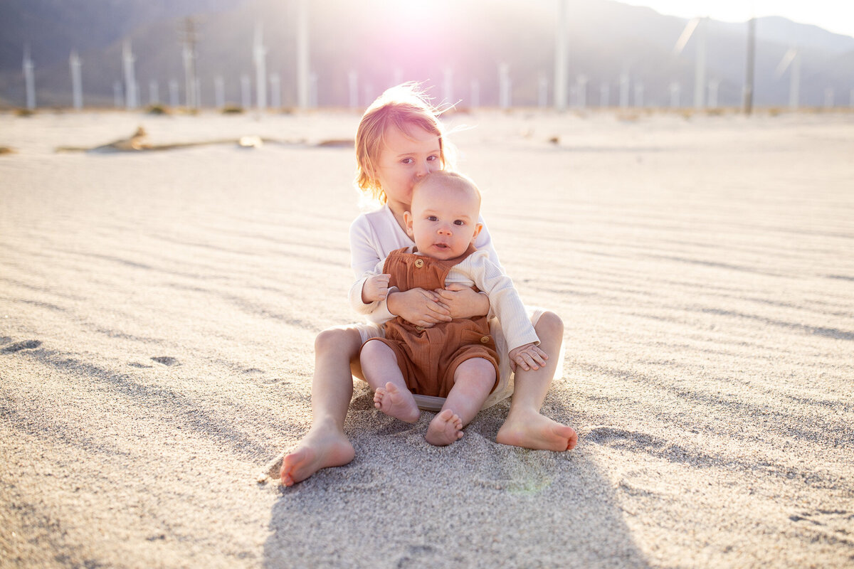 Siblings sit in the sand in front of large windmills at sunset. 
