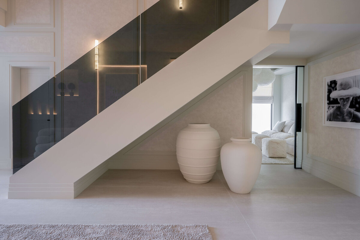 A side view of a large modern staircase,bordered by a sleek, black glass railing. Beneath the staircase are two large cream-colored vases.