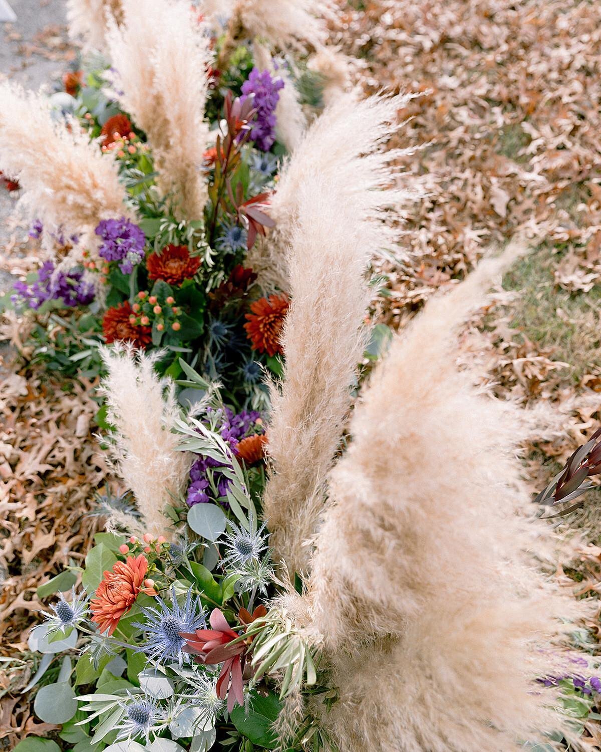 A long floral arrangement sits on the floor leading up to the ceremony arbor. The floral centerpiece features pampas grass, blue thistle, silver dollar eucalyptus, red mums, orange mums, purple flowers and yellow flowers.