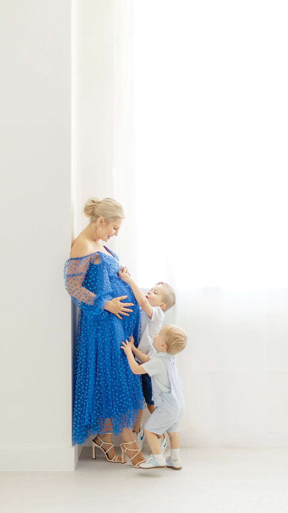 A pregnant mother standing against a wall in a Flower Mound photography studio while her little boys are embracing moms growing belly.