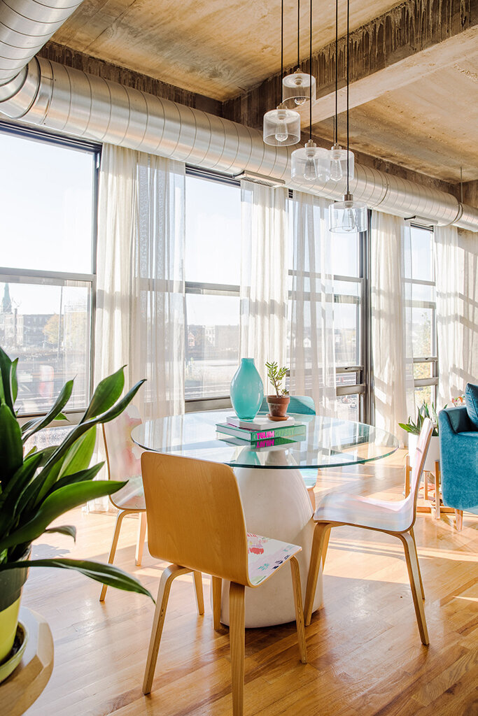 Modern and eclectic dining space with a glass-topped table and minimalist wooden chairs. The room features large windows with sheer curtains, exposed ductwork on the ceiling, and warm wooden floors. A pop of turquoise is provided by a vase on the table.