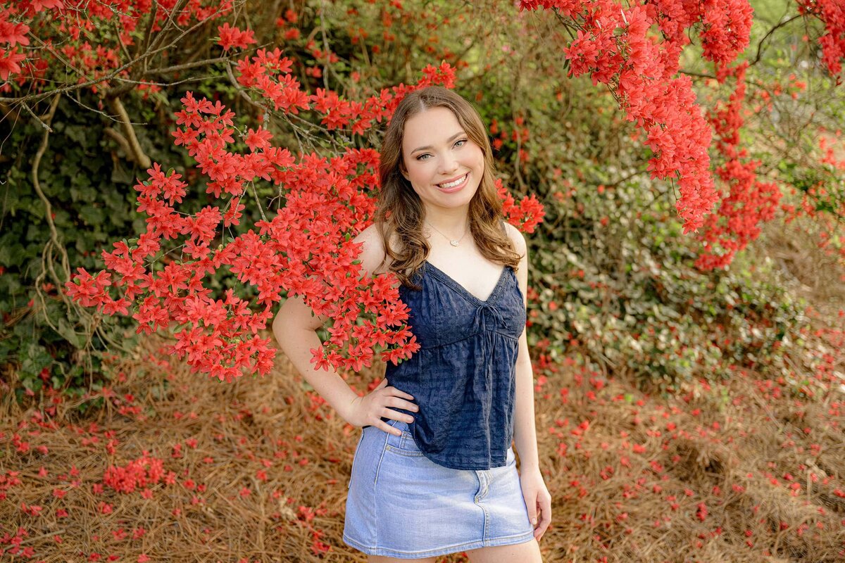 charlotte area senior stands in blue shirt and jean skirt in a rock hill garden surrounded by red blooms for her senior photos