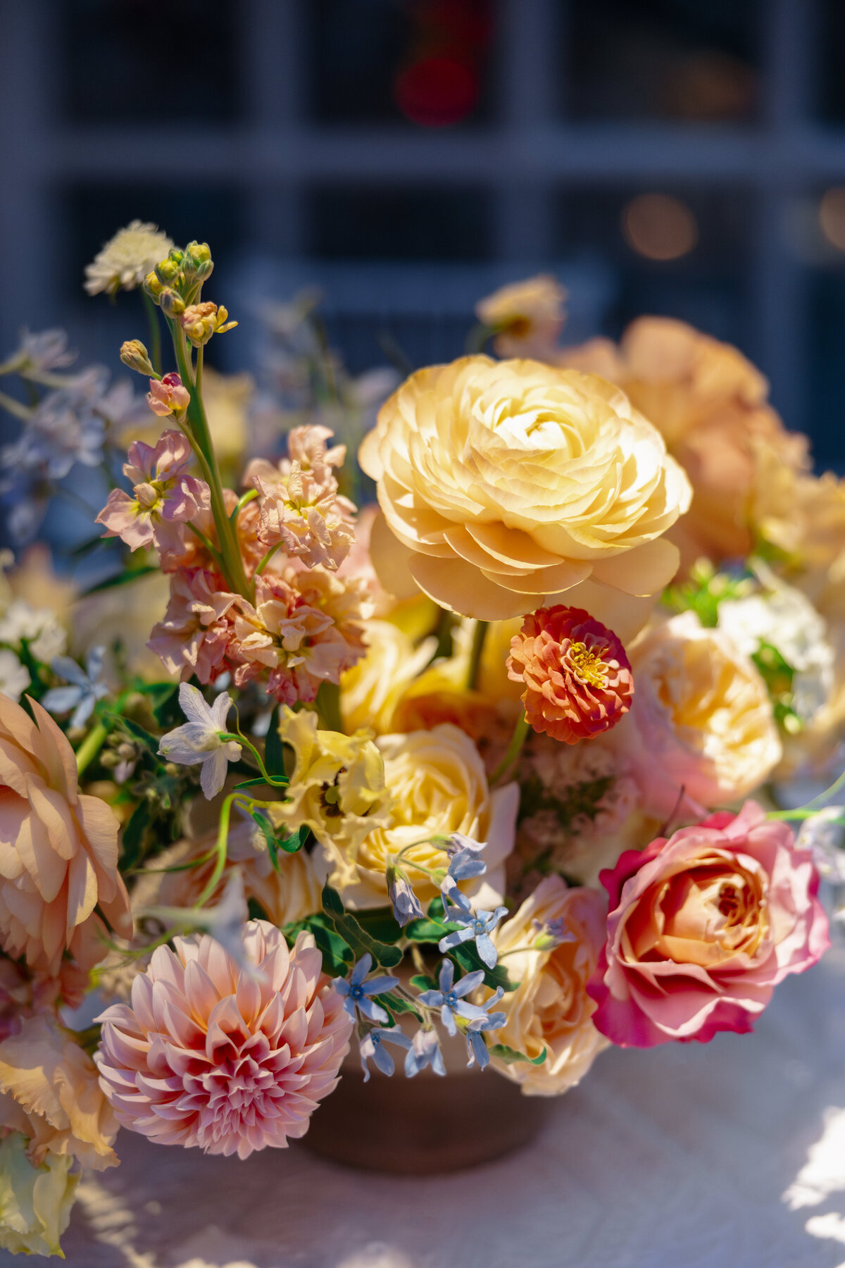 A vibrant floral arrangement featuring an assortment of flowers in shades of yellow, pink, and peach. The bouquet sits on a table, showcasing roses, dahlias, and other blooms, softly illuminated against a blurred dark background.