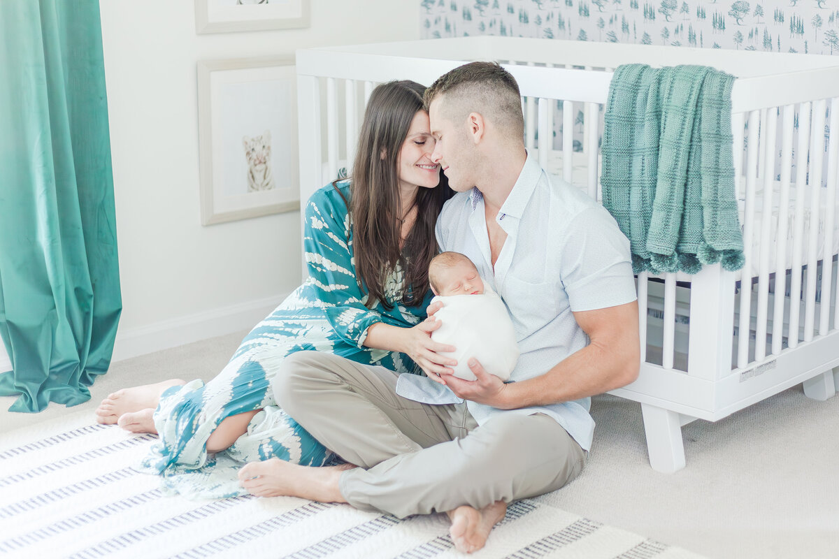 Mom and dad sitting on floor of nursery with dad holding newborn baby boy