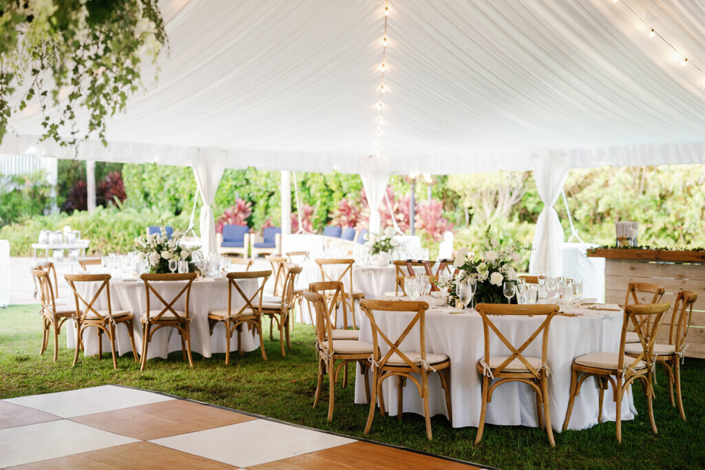A beautifully styled outdoor wedding reception with soft white draping, rustic wooden chairs, and elegant floral arrangements, set against a lush tropical backdrop. Captured by Claudia Amalia Photography.