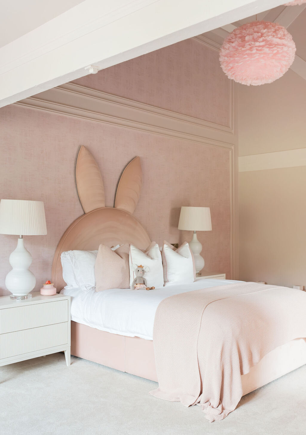 A cozy girl’s bedroom features a soft pink color palette, with a bed adorned with a unique headboard shaped like bunny ears. Two modern white nightstands with large white lamps flank the bed. The bedding is light pink, and a fluffy pink chandelier hangs overhead.