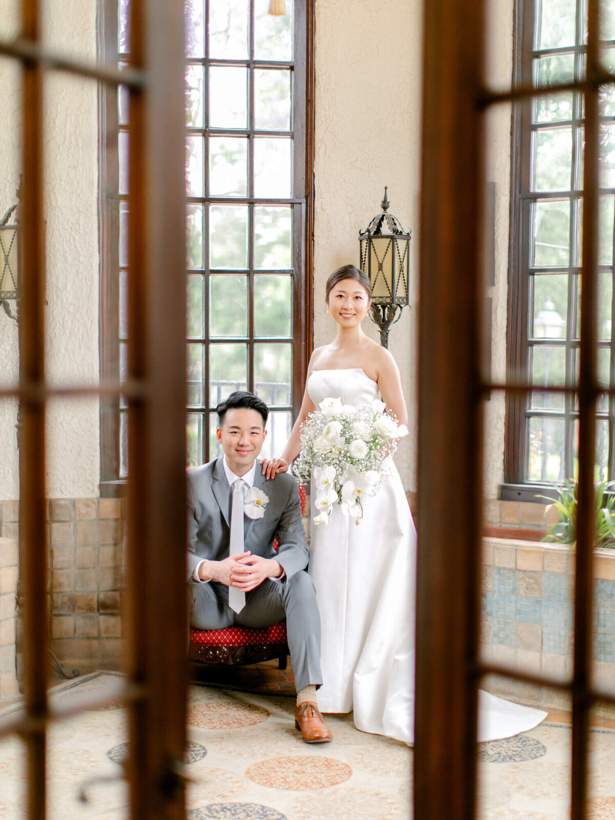Bride in a white dress and groom in a gray suit pose indoors by tall windows, framed by wooden door panels. The bride holds a bouquet, both smiling warmly.