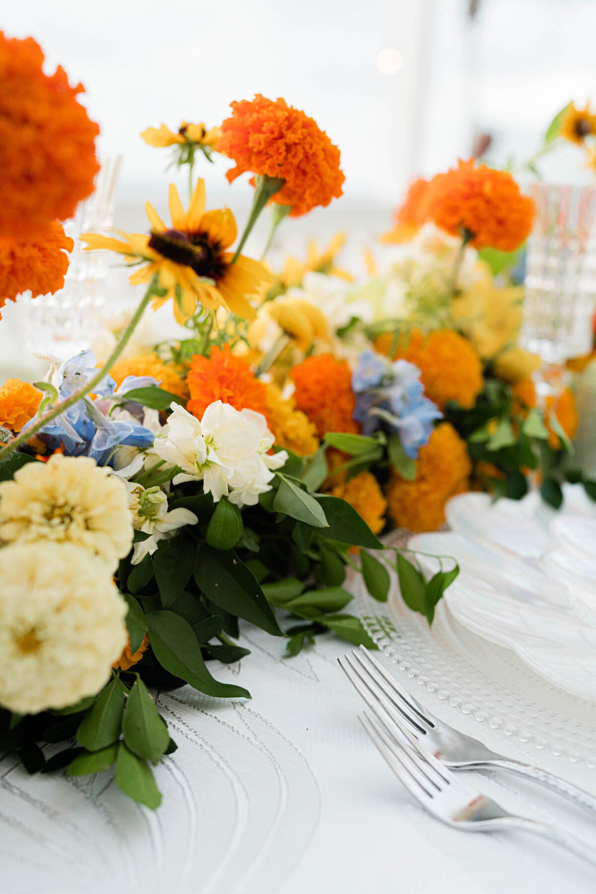 white on white palm leaf tablecloth with hammered silver flatware and an iridescent seashell plate adorned with a white, orange, blue and green floral table runner
