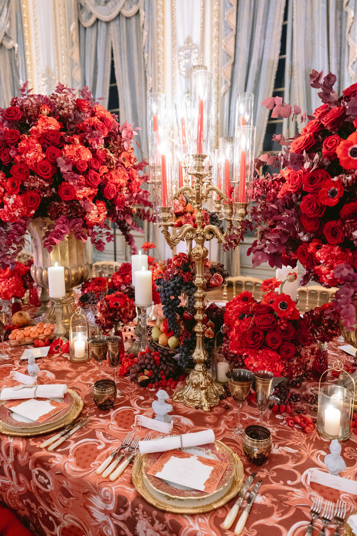 The lavish dining table, beautifully captured by photographer Matteo Coltro and videographers Zen Film Works, was set at the exquisite Shangri La Hotel & Ritz Paris Hotel in Paris, France. The scene is simply breathtaking, with an ornate display of red roses, candles, and gold accents adorning the table. Tall candelabras with striking red candles stand elegantly amidst the intricately designed tableware embellished with gold trim, all set against the backdrop of large windows draped with luxurious curtains. It truly is a scene of opulence and sophistication.