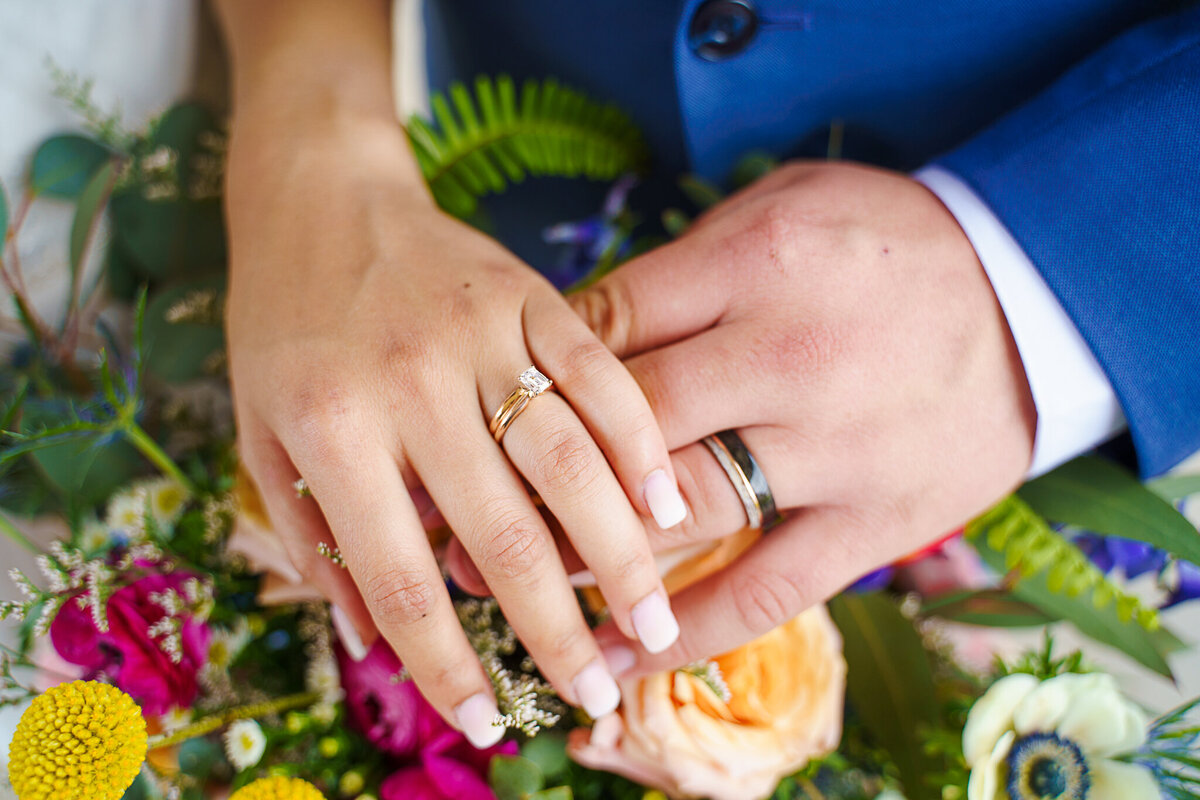 A bride and groom put their hands together over a bouquet to show off their rings.