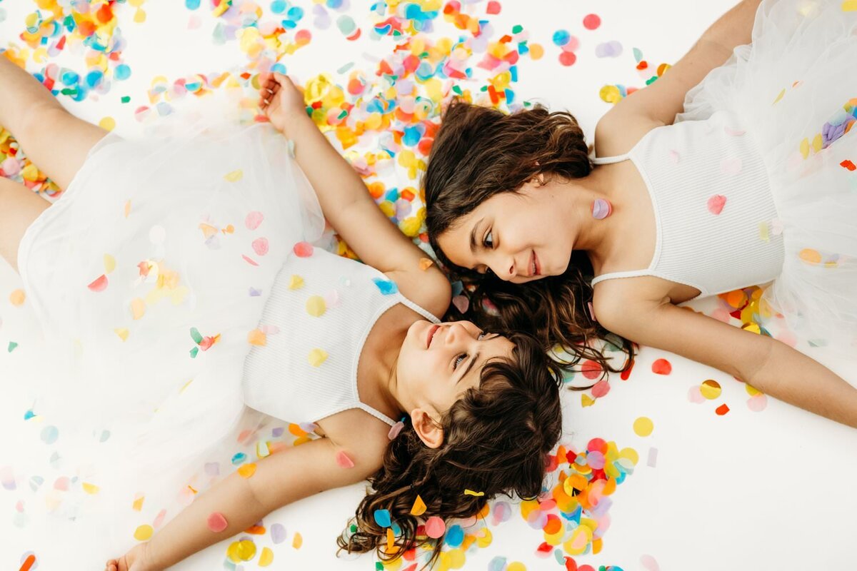 sisters surrounded by confetti in a photography studio. they look at each other on a white backdrop