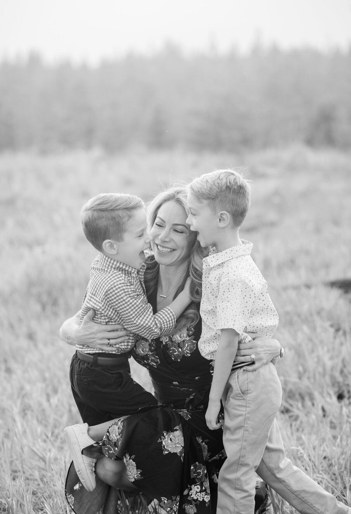 This black and white photo shows a mom kneeling down to the level of her young sons. They are all laughing and smiling with each other