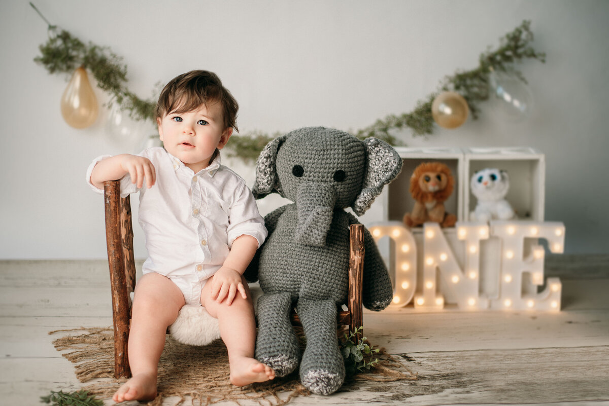 One year old brown haired boy wearing white button down shirt sitting on small wooden bed with knitted stuffed elephant and festive balloons and animals in the backdround