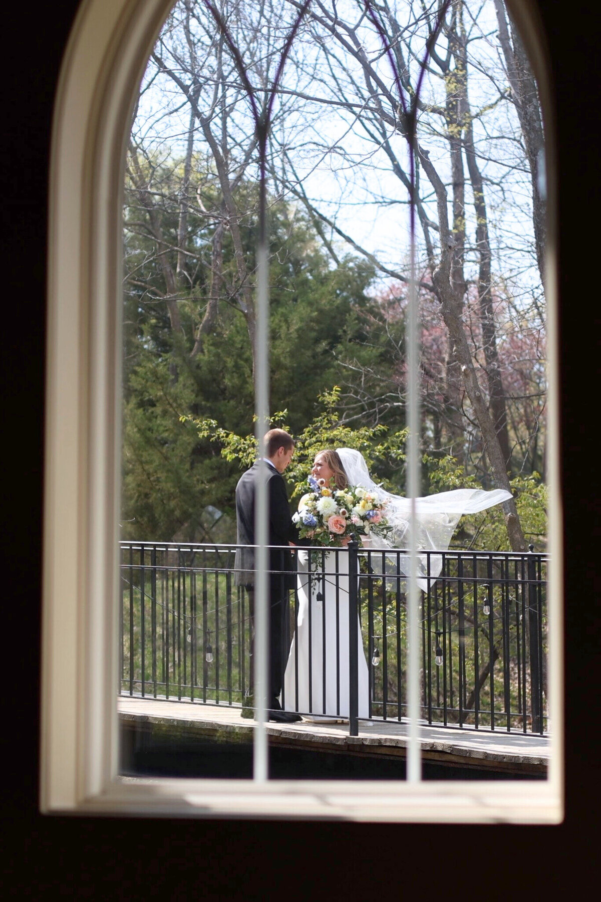 Bride and Groom on bridge with bride's veil blowing in wind captured through church window at Hawthorne House