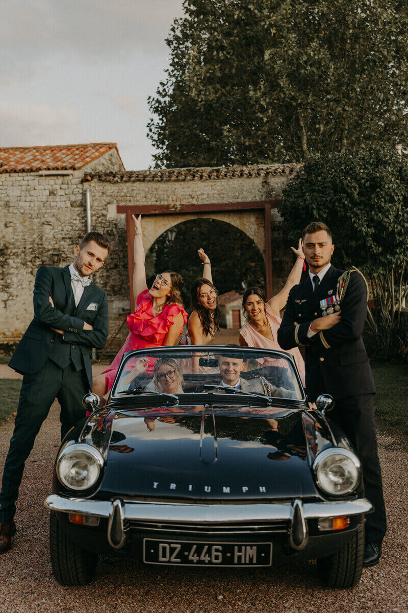 Mariés assis dans une voiture vintage noire,  amis autour prenant la pose pour Laura, photographe professionnelle en Vendée.