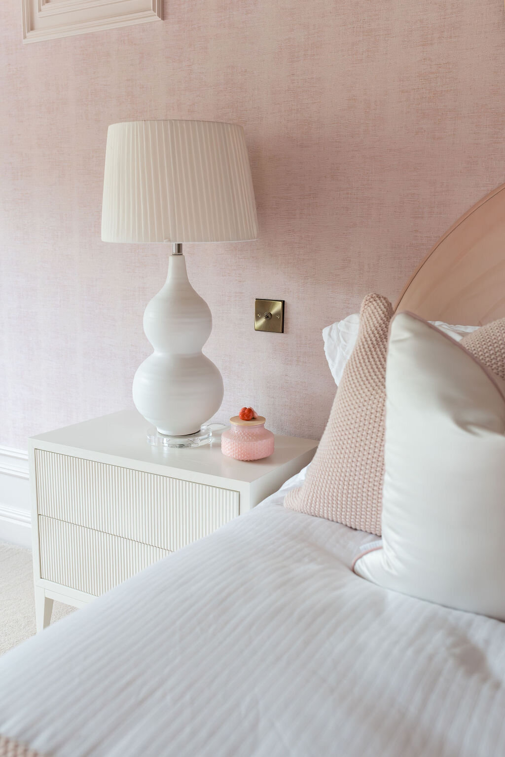 A modern white nightstand with a large white lamp beside a girl's pink bed and soft pink walls.