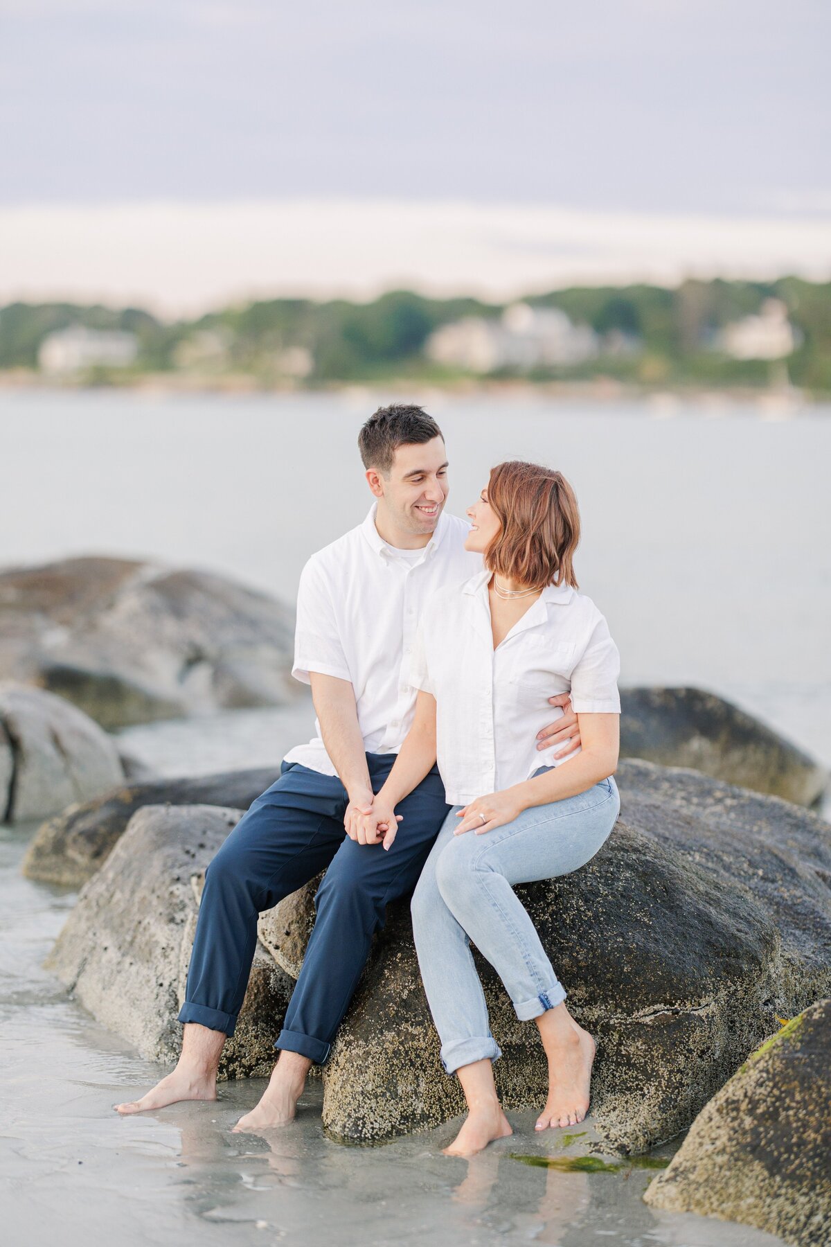 A couple holding hands and smiling at each other during their sunset Wingaersheek Beach engagement photos.