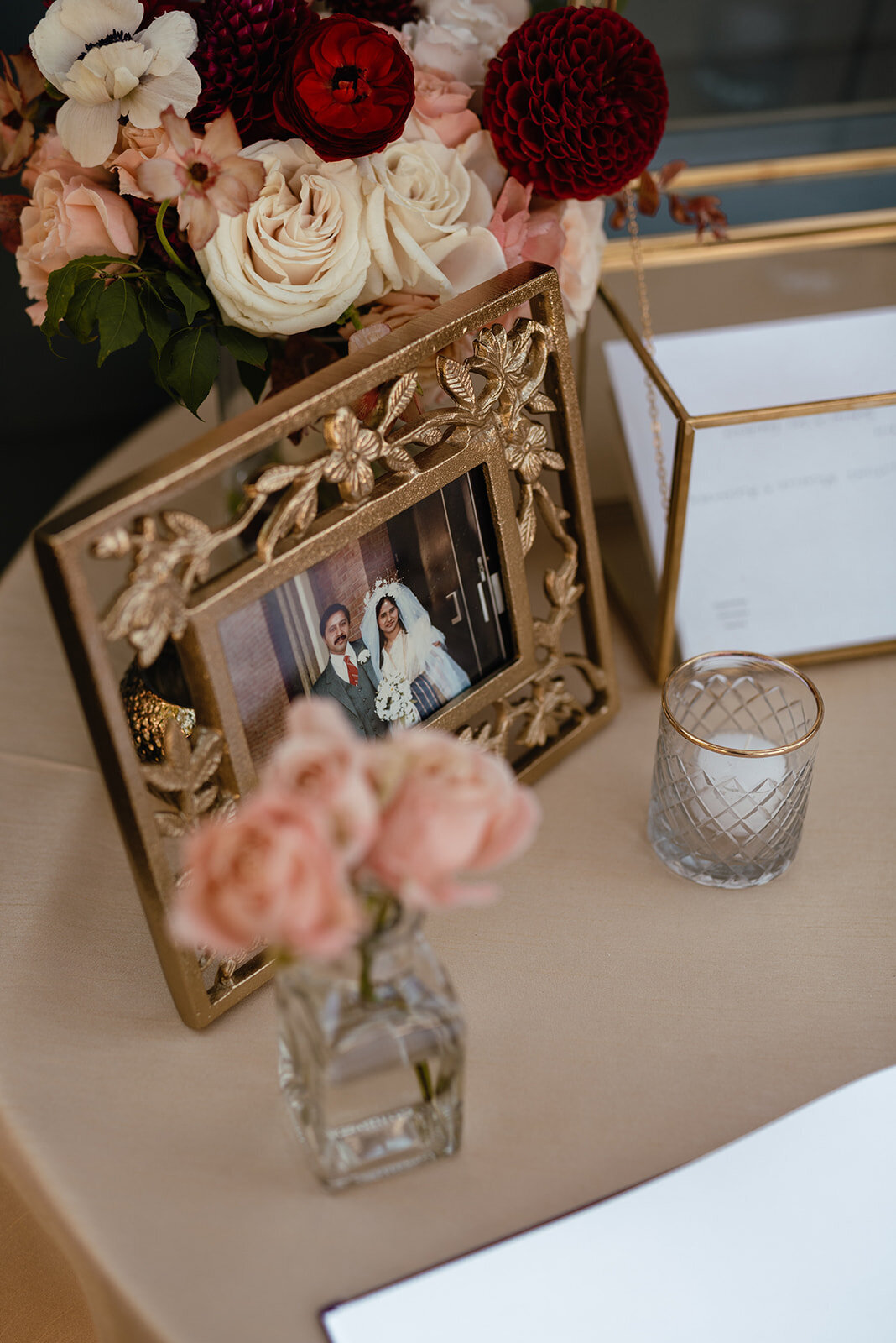 A beautifully decorated guestbook table with an ornate gold frame displaying a couple’s portrait, soft pink florals in glass vases, and a candle in a textured holder.