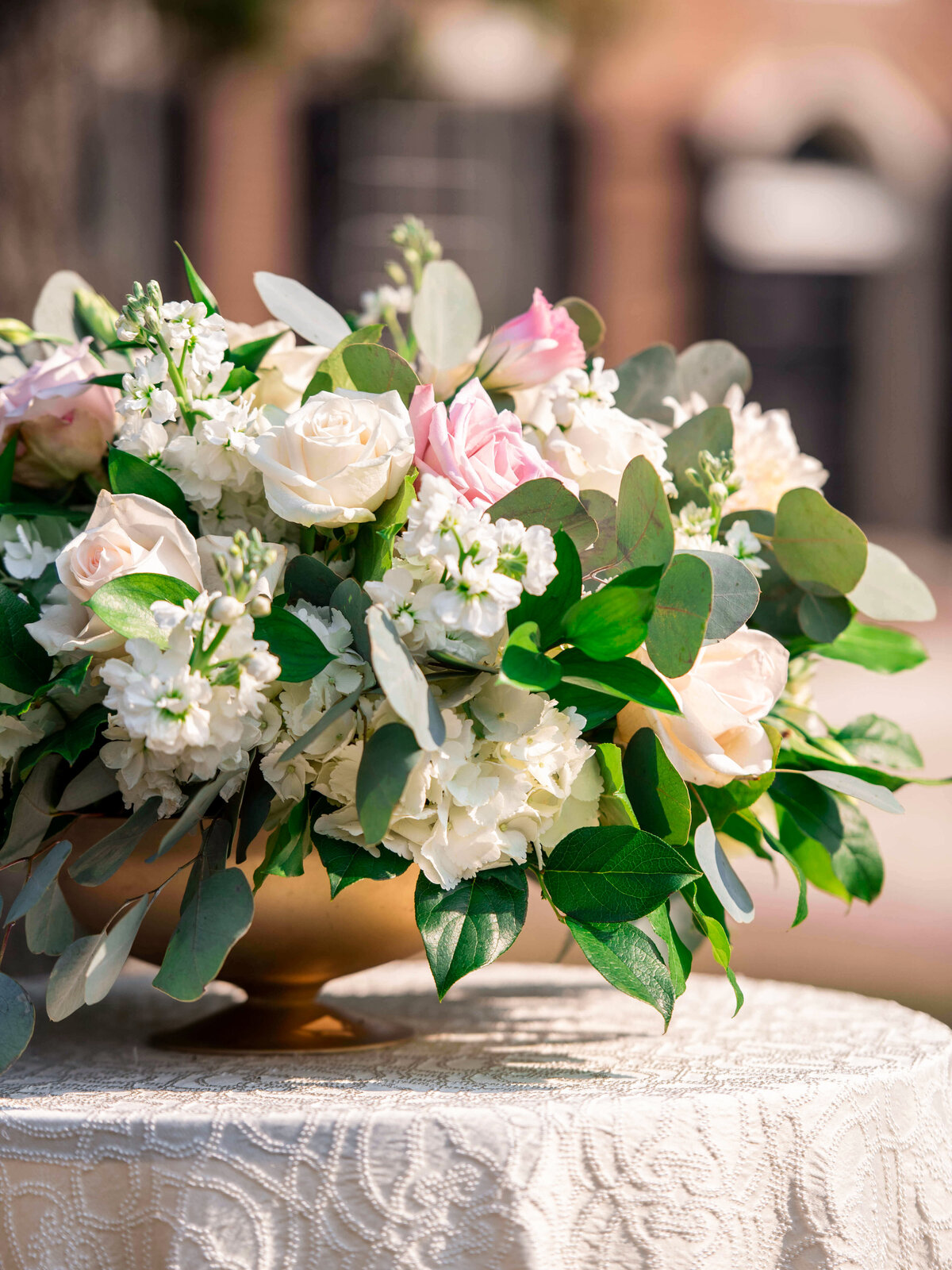 A floral centerpiece with white roses, pink tulips, and lush greenery in a wooden bowl sits on a lace-covered table. The background is softly blurred, suggesting an outdoor setting.