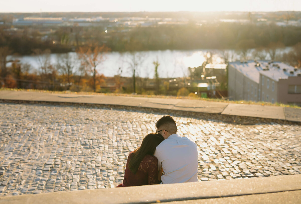 fall engagement photos with Richmond wedding photographer capturing man and woman cuddling on a bench at golden hour with the sun setting over the horizon of the city