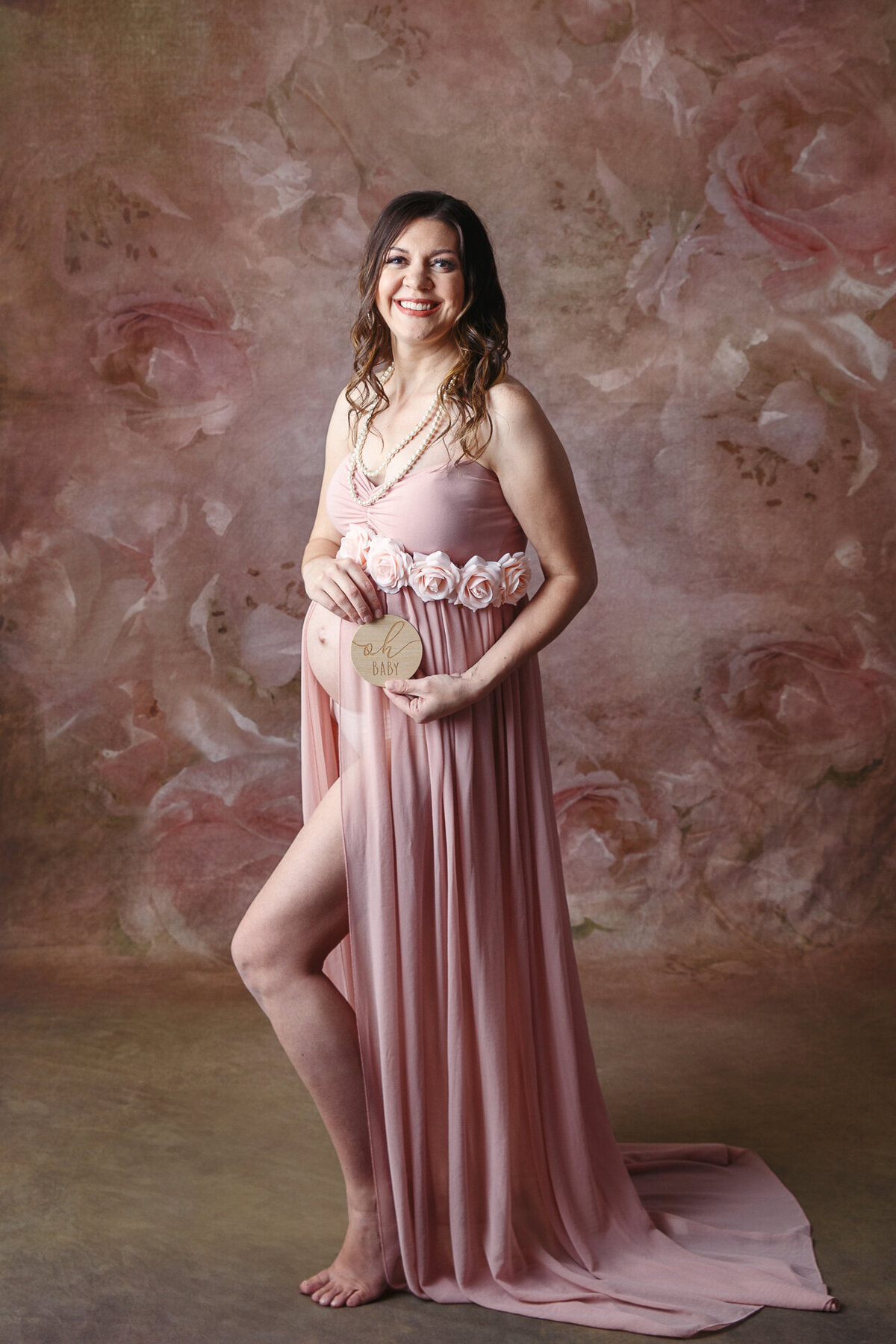 Pretty woman wearing a pink maternity dress and holding a oh baby sign smiling