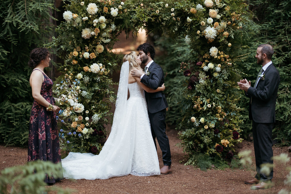 The bride and groom's first kiss in front of their ceremony arch, with their wedding party looking on