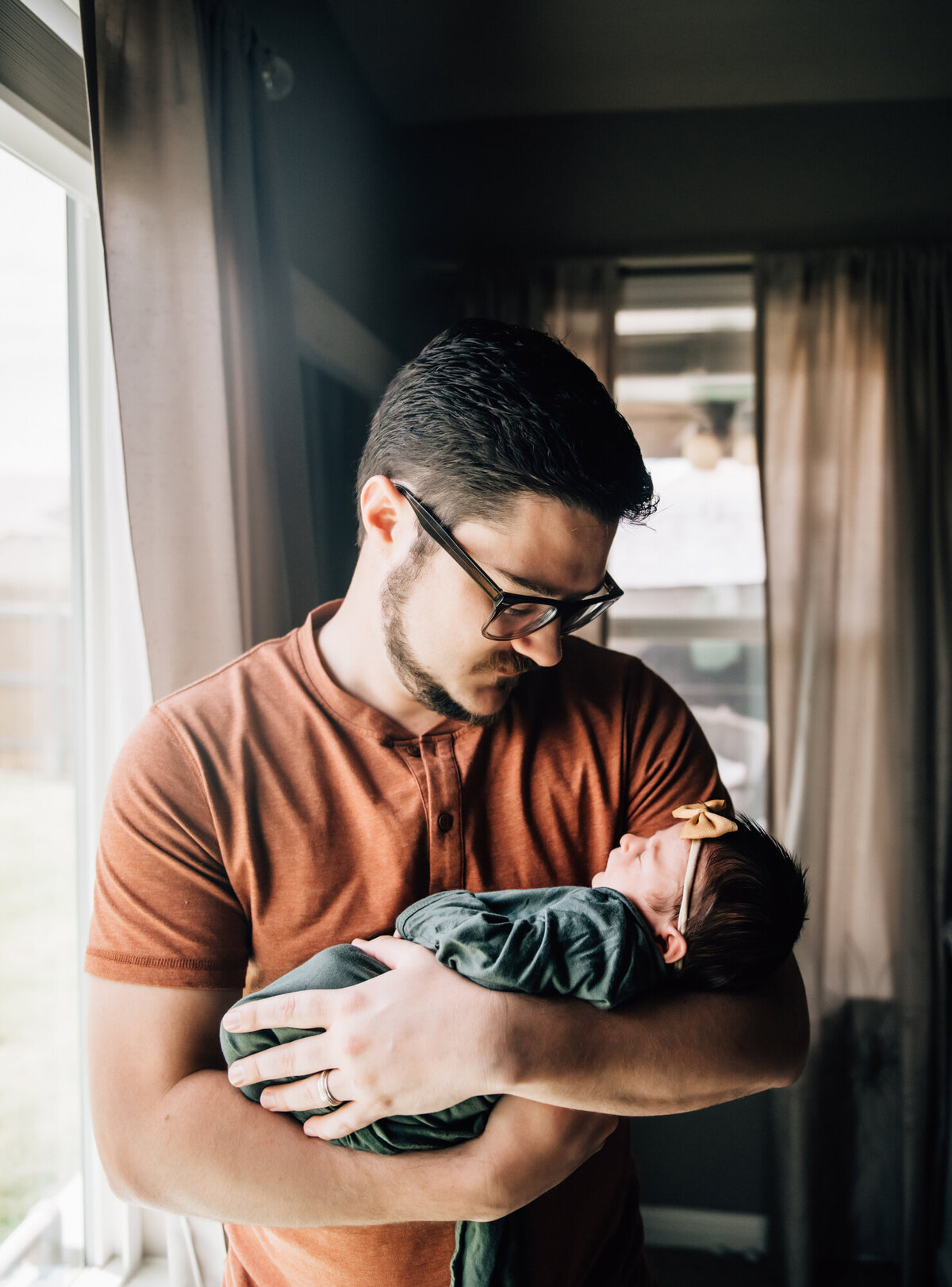 Newborn Photographer, Dad is holding baby girl and smiling at her while standing by a window.