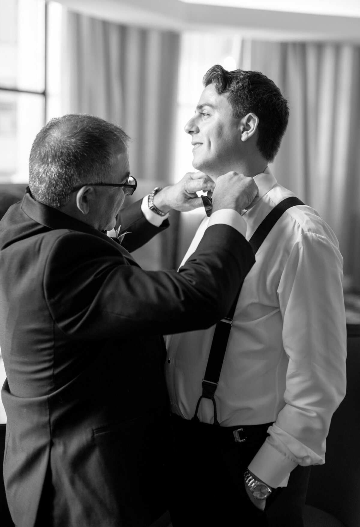 A man in a suit adjusts the bow tie of another man who is wearing suspenders and a white shirt. They are both smiling, standing in a room with curtains in the background.