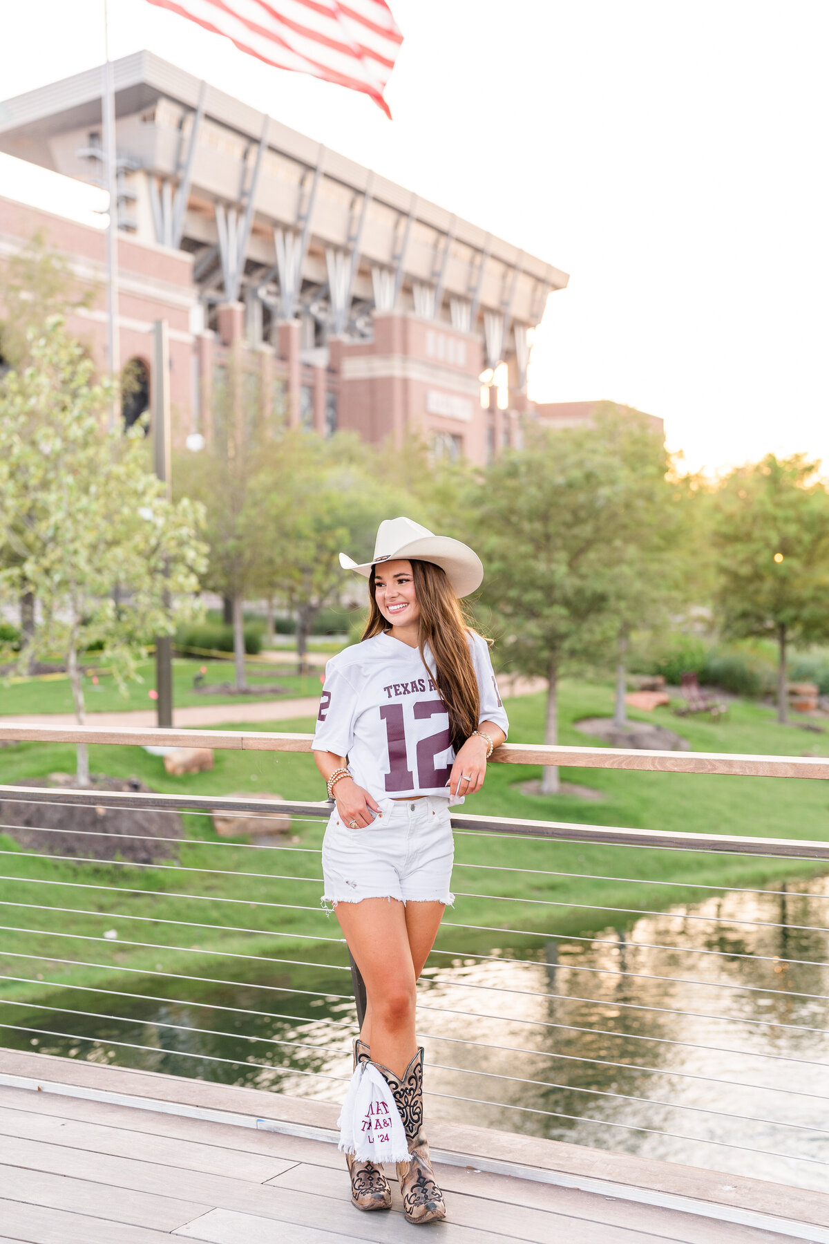 Texas A&M senior girl leaning on bridge railing with other hand in pocket while wearing white jersey, shorts and cowboy hat and smiling away in front of Kyle Field in Aggie Park