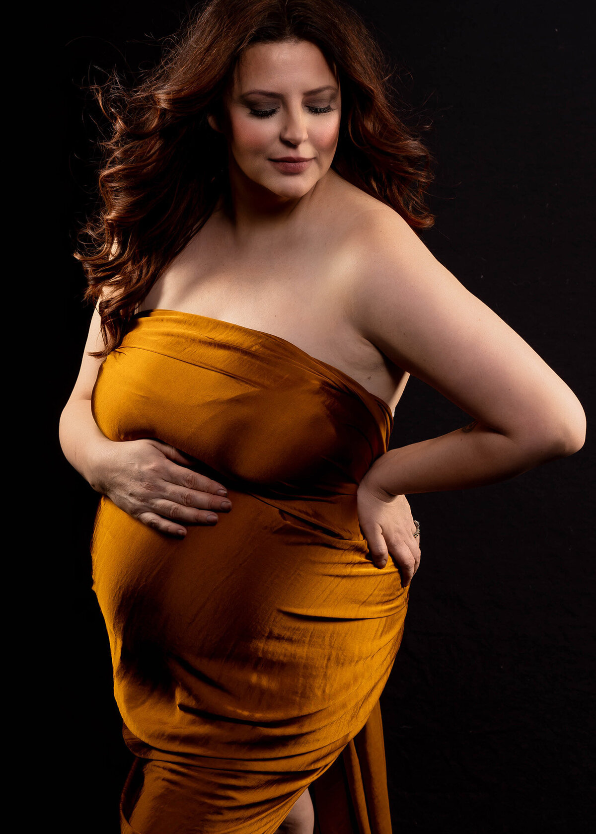 A pregnant woman wrapped in gold fabric stands in a studio with hands on hip and bump and looking down shoulder in the studio of an Iowa maternity photographer