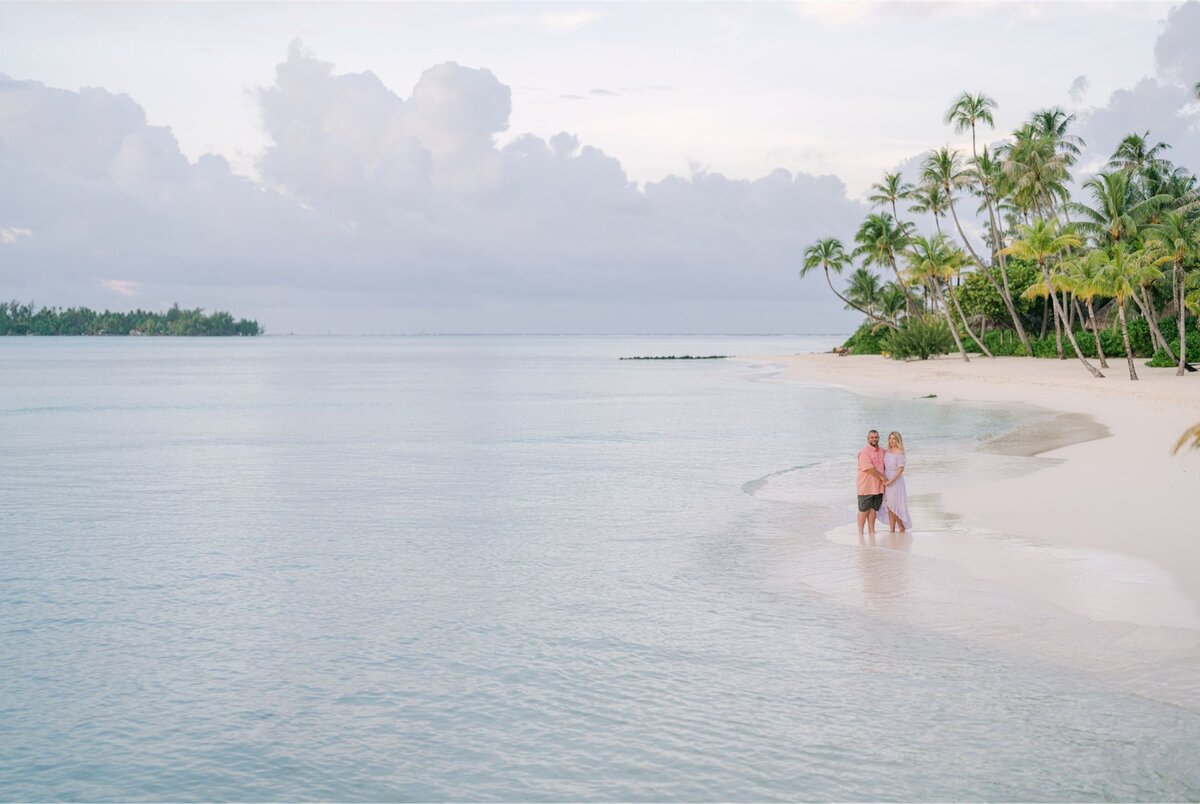 Couple walking along a white sand beach at sunset, surrounded by calm turquoise waters and palm trees, enjoying a luxury tropical vacation.