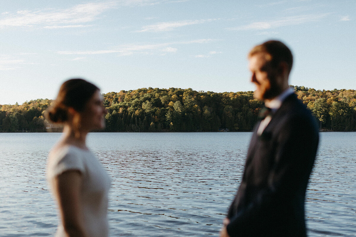 A couple stands facing each other by a serene lake, captured by a talented Toronto wedding photographer. Blurred figures grace the foreground while majestic trees and an open sky form a picturesque backdrop.