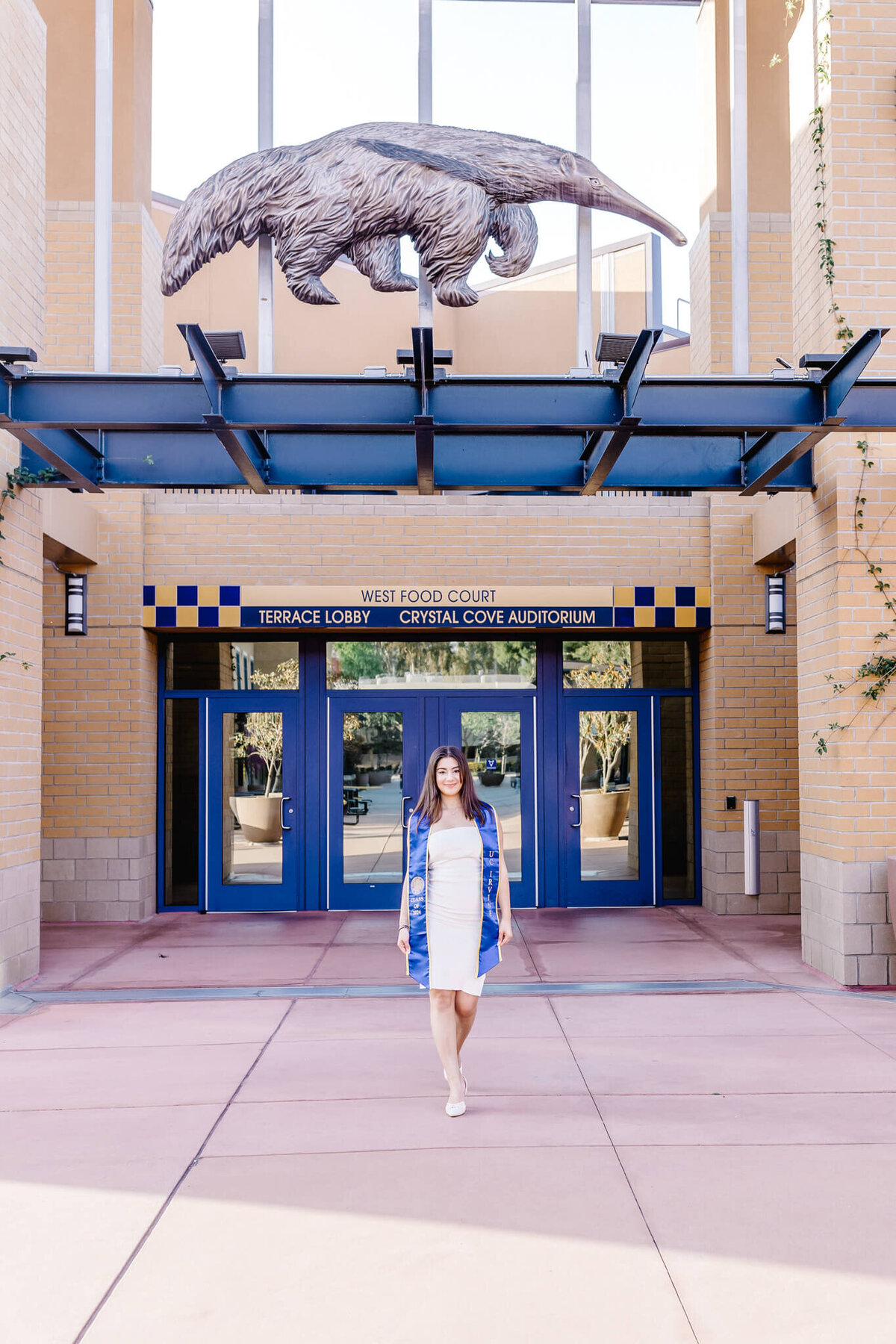 A person in a white dress and blue jacket walks in front of a building with blue doors. Above is a large sculpted figure of an animal on a beam. The building has signs reading Terrace Lobby and Crystal Cove Auditorium.