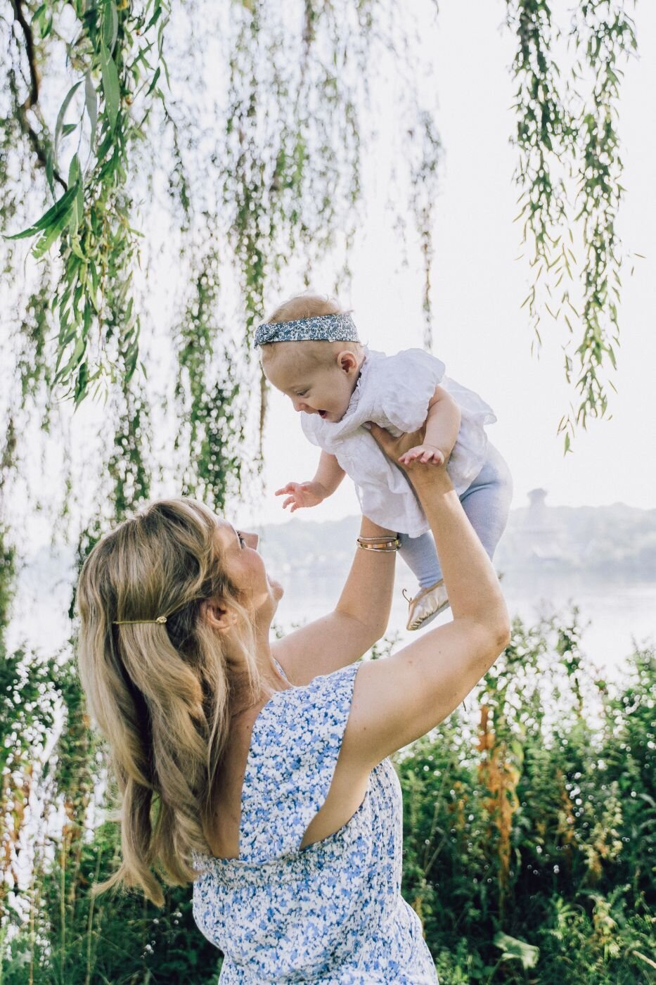 Mother lifting smiling baby girl wearing floral headband under willow trees by the water.