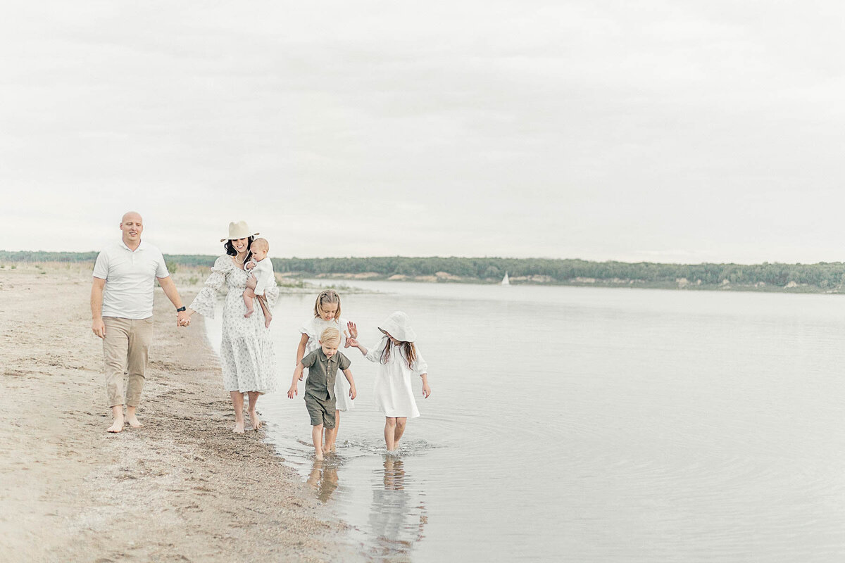 A family of six holds hands and walks by the lake during a beach photo session in Kansas City to celebrate their baby’s 6-month milestone.