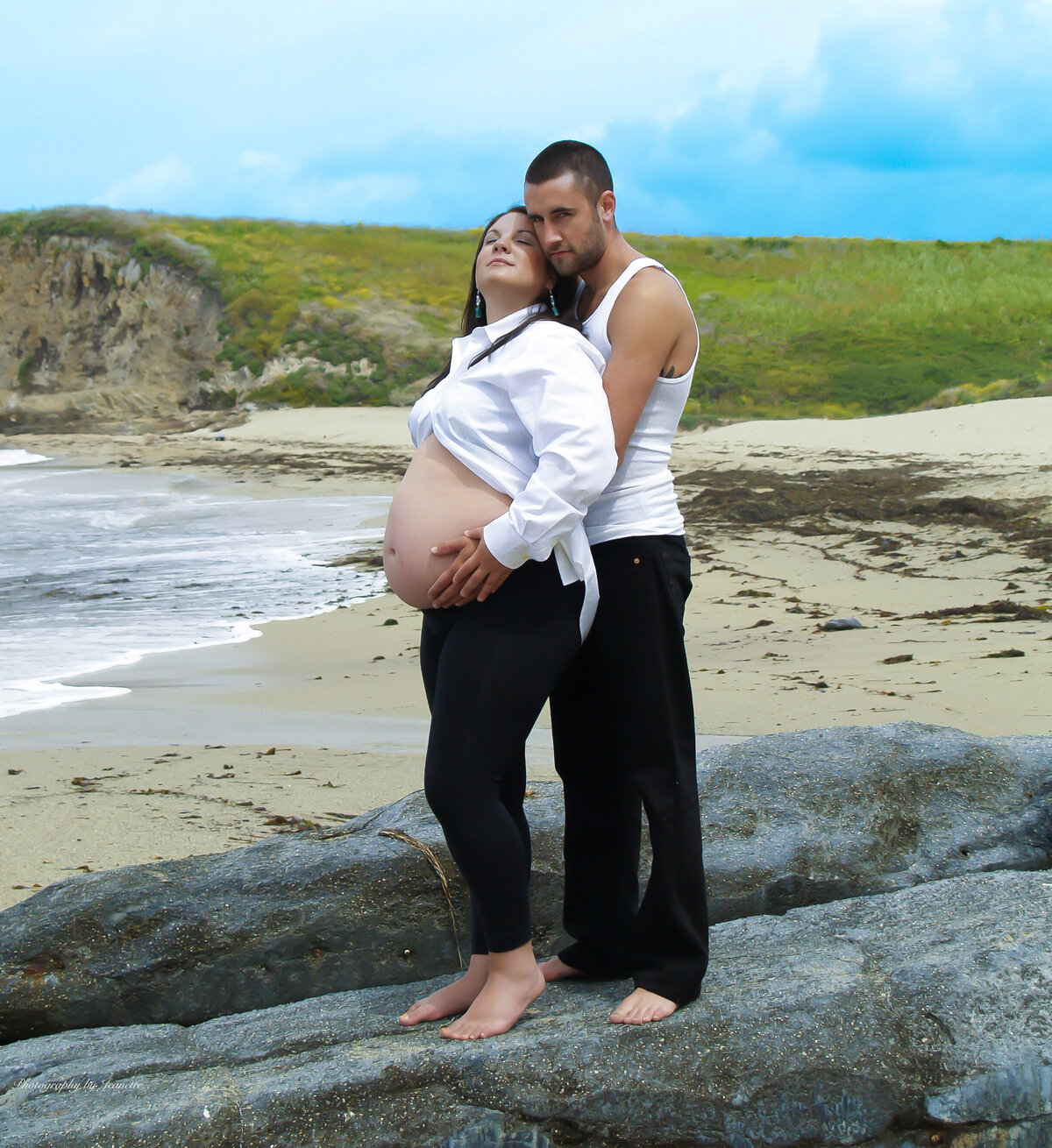Husband hugging his Pregnant wife while standing on rock at beach with beautiful green grass and turquoise sky background