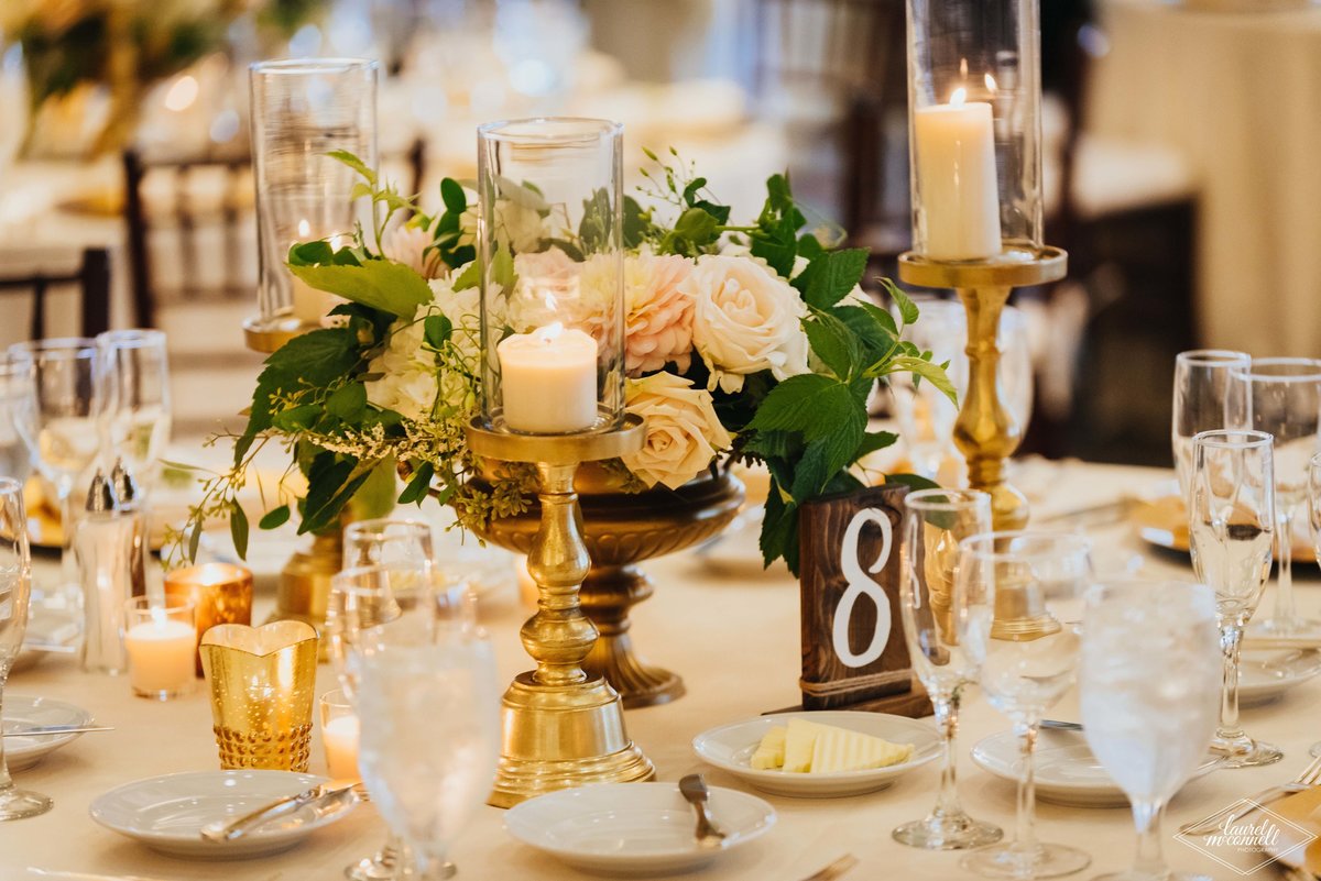 gold compote centerpiece of peach and ivory flowers with greenery, wooden table number sign, gold candle holders,and gold mercury votive candles