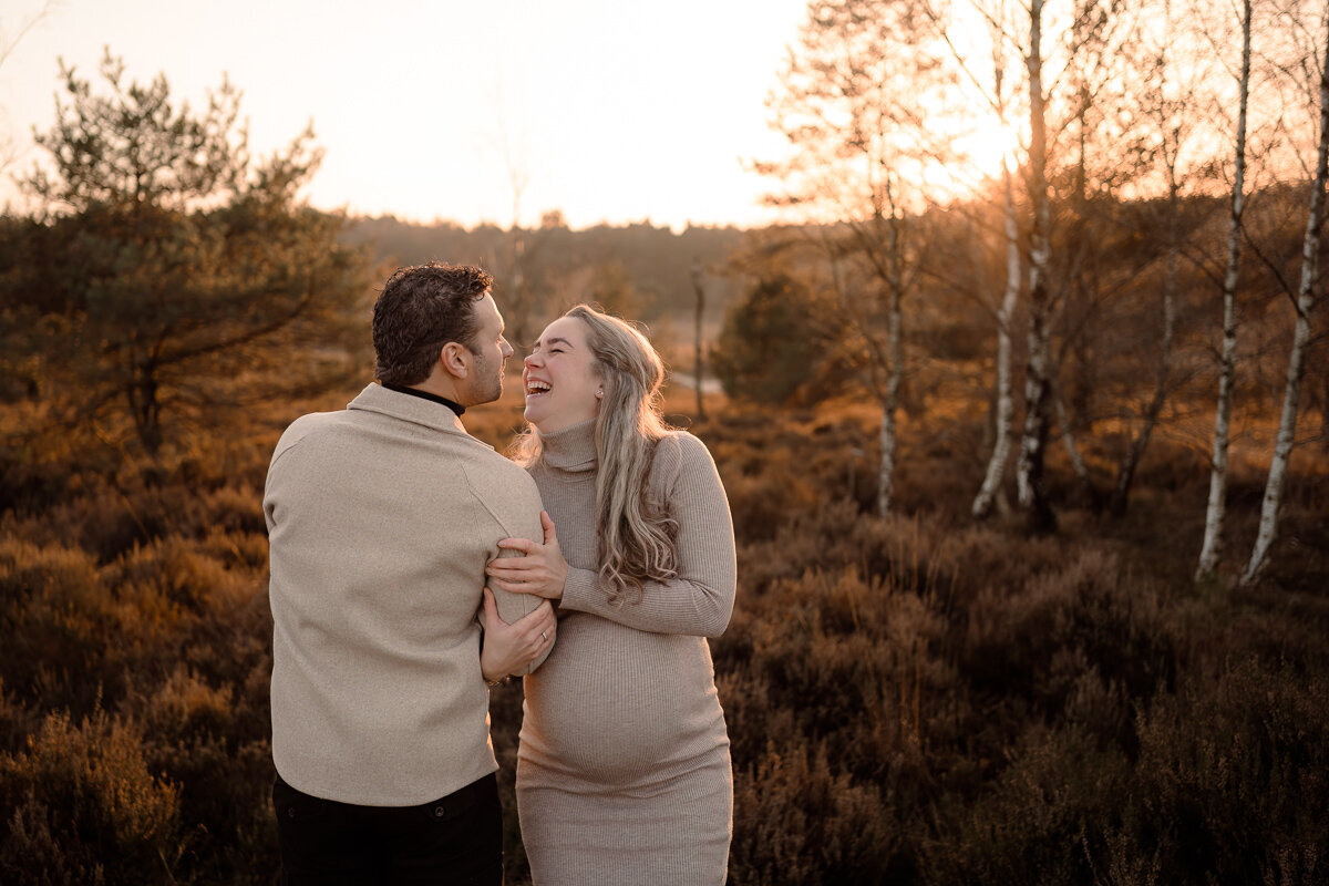 Zwangere vrouw staat naast haar man met haar armen ingehaakt in de zijne en ze kijken elkaar lachend aan