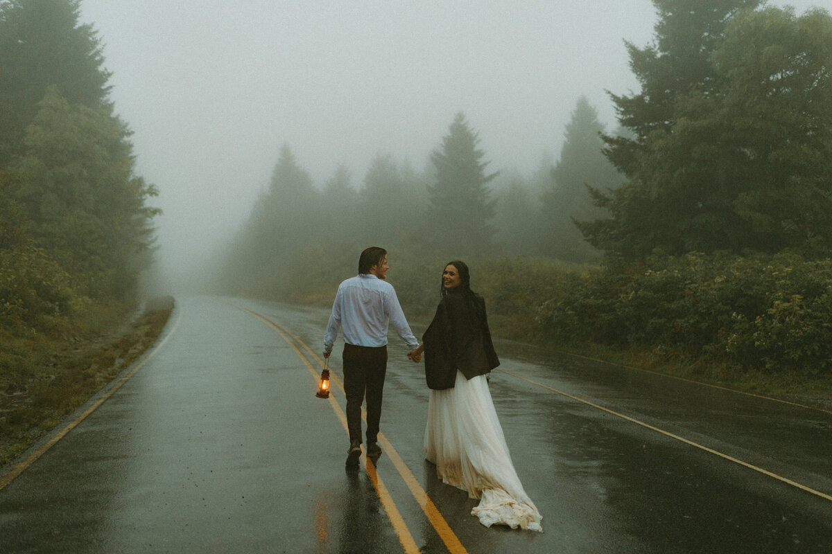 bride and groom holding lanterns walking in rain on a road  after elopement on mountain