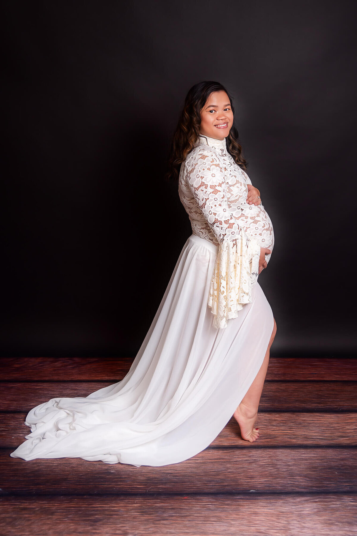 A pregnant woman in a flowing white dress smiles as she poses for a photo, showcasing her beautiful baby bump.